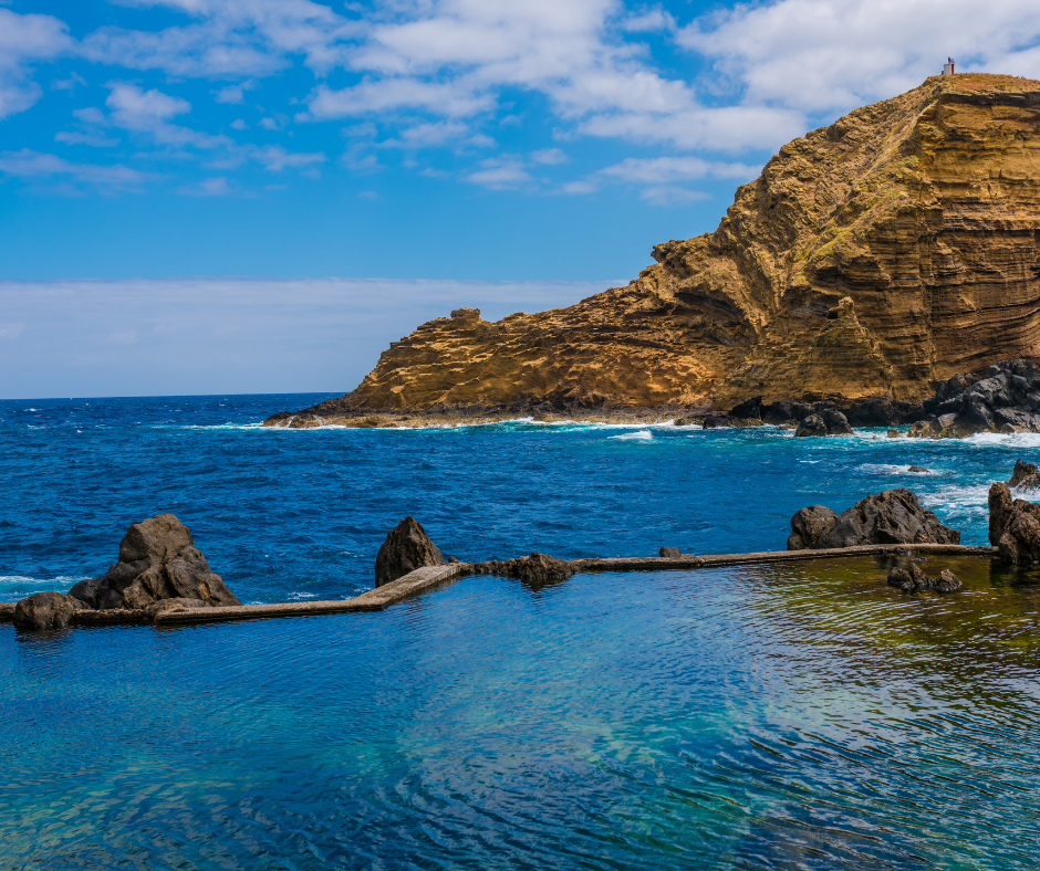 Scenic coastal view of Porto Moniz on Madeira Island. Rocky cliffs surround natural volcanic pools filled with clear blue ocean water. Waves crash against the rocks while visitors relax in the pools and enjoy the breathtaking natural beauty