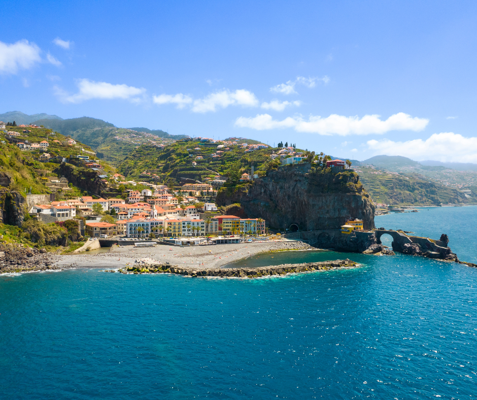 "Spectacular view of Ponta do Sol on Madeira Island. The picturesque coastal village is bathed in golden sunlight, with colorful buildings nestled between lush mountains and the sparkling azure ocean. Waves gently caress the rocky shoreline, creating a serene and inviting scene